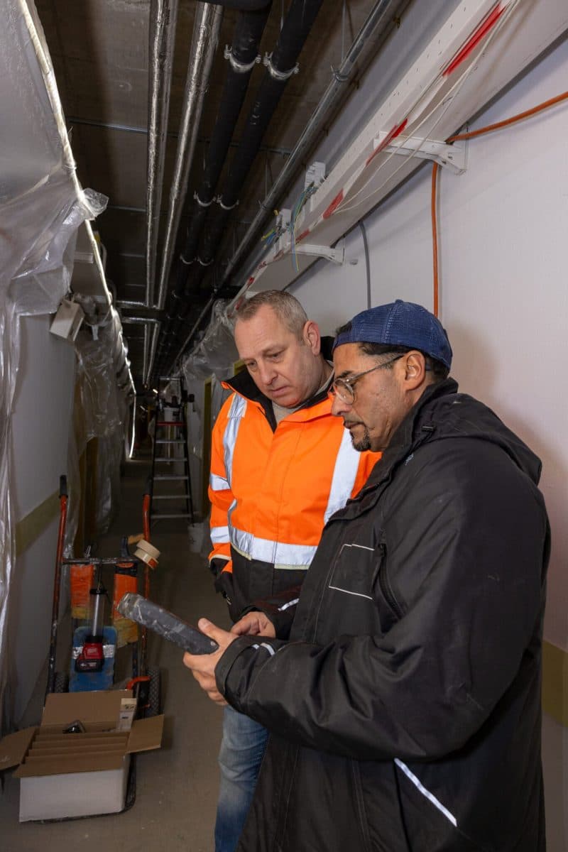Twee monteurs van WCC bekijken het digitale logboek op een tablet, in een gang met leidingen